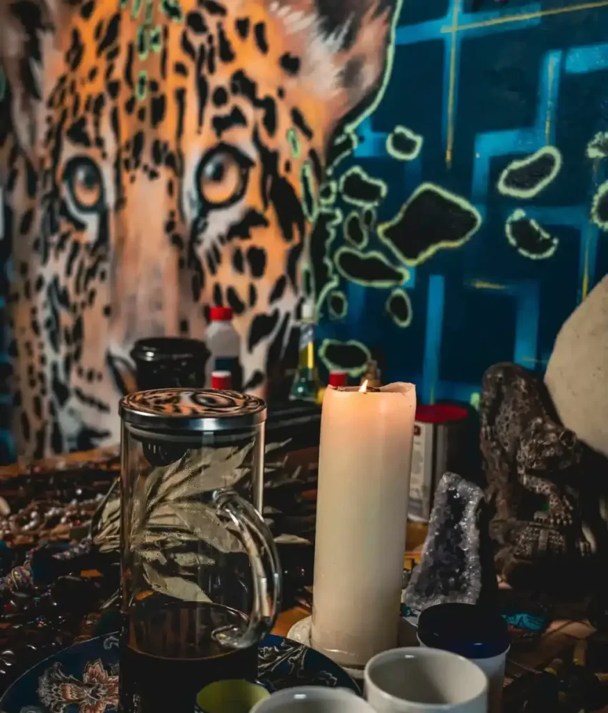 ceremonial altar with the candle, ayahuasca medicine, cups, cascavelles, the waira and more atop the altar. Artwork of the jaguar can be seen in the background.
